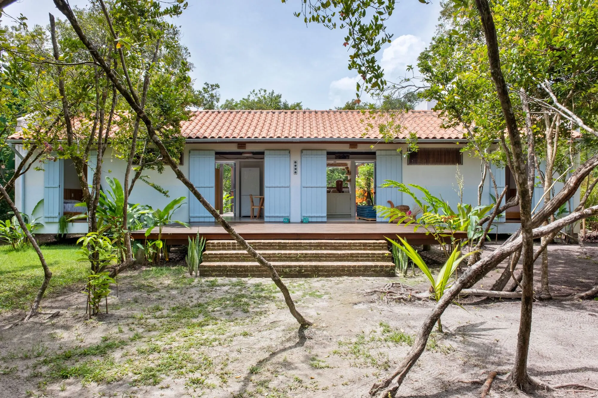 Fachada azul da Casa Palco vista entre os troncos, com o deck e a escada de acesso